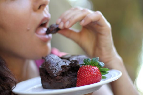 Woman eating a piece of a brownie while holding a plate carrying a strawberry and brownie