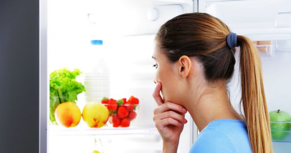 woman with a ponytail thinking while looking at an open fridge filled with fruits and vegetables