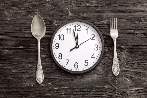 A clock in between a fork and a knife on a grey, wooden table.